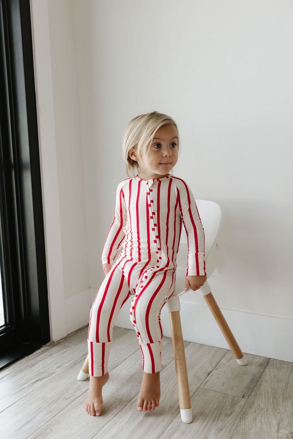 A child with blonde hair sits on a white chair by the window, dressed in breathable Bamboo Zip Pajamas from forever french baby—a onesie featuring red vertical stripes. The room resembles a serene sanctuary, boasting light wooden floors and pristine white walls.