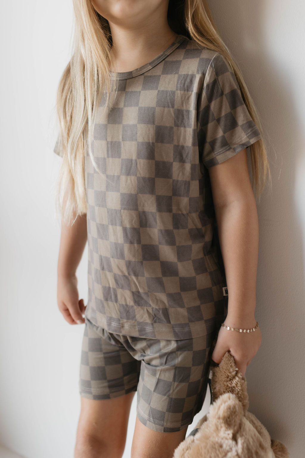 A child with long blonde hair wearing hypo-allergenic clothing—the Bamboo Short Set in a Faded Brown Checker pattern by forever french baby—stands against a white wall. The child holds a stuffed animal close, their face not visible in the image.