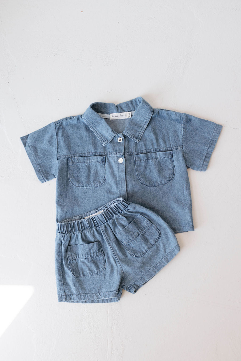 A neatly laid out Denim Short Set | Cash from Forever French Baby, consisting of a light denim short-sleeved shirt with buttons and two front pockets, paired with matching light denim shorts that have an elastic waistband and two front pockets. This adorable two-piece denim set is arranged on a light background.