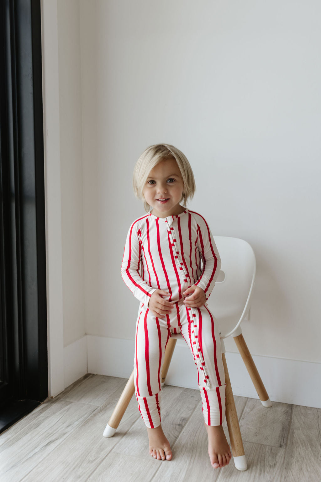 A child with blonde hair sits barefoot on a white chair in a room with light wood flooring, smiling at the camera. They are dressed in the Bamboo Zip Pajamas | the Claus from forever french baby, radiating comfort and charm with its breathable fabric featuring red vertical stripes and buttons.