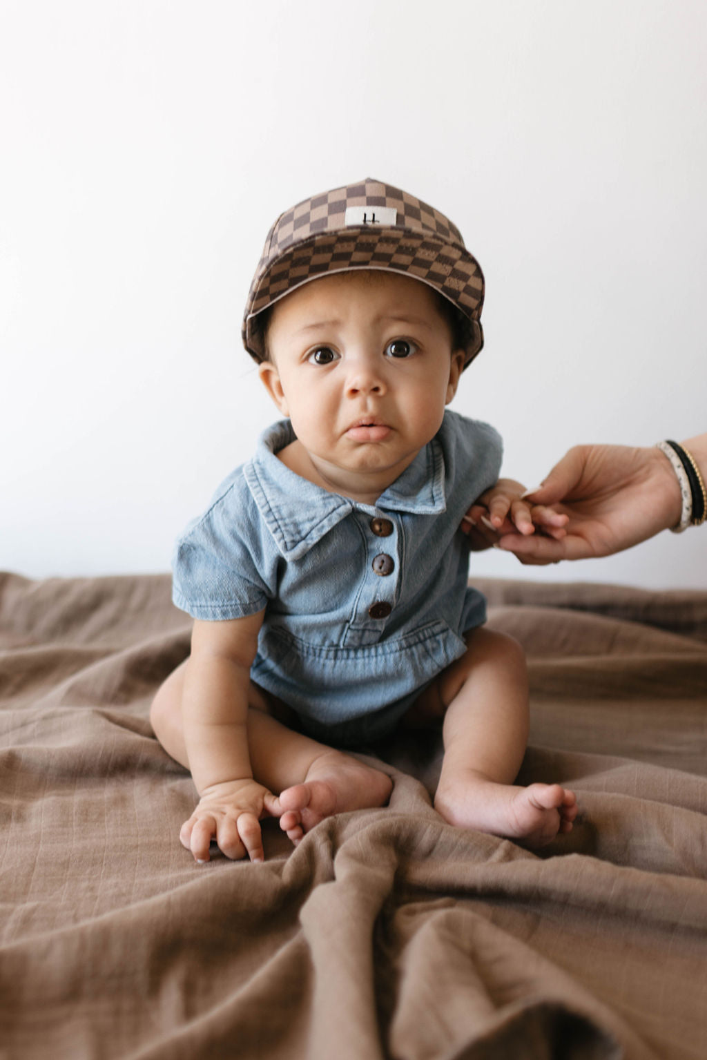 A seated baby, sporting a checkered cap and a stylish Denim Romper from forever french baby with buttons, is gently supported by an adult's hand. The baby has a curious expression and sits on a brown blanket against a plain background, epitomizing Forever French Baby fashion.