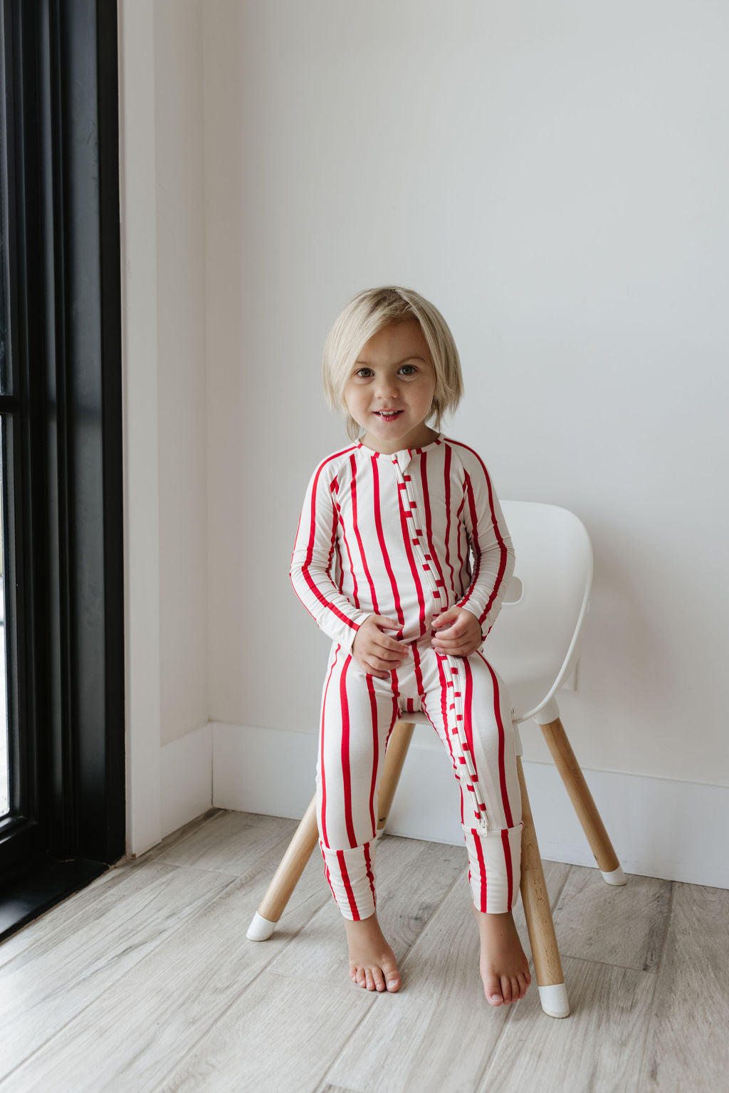 A young child with short blond hair sits on a white chair near a window, wearing the Bamboo Zip Pajamas in the Claus pattern from forever french baby, featuring breathable red and white stripes. Smiling at the camera, they enjoy the comfort of their pajamas. The room has light wooden flooring and a plain white wall.