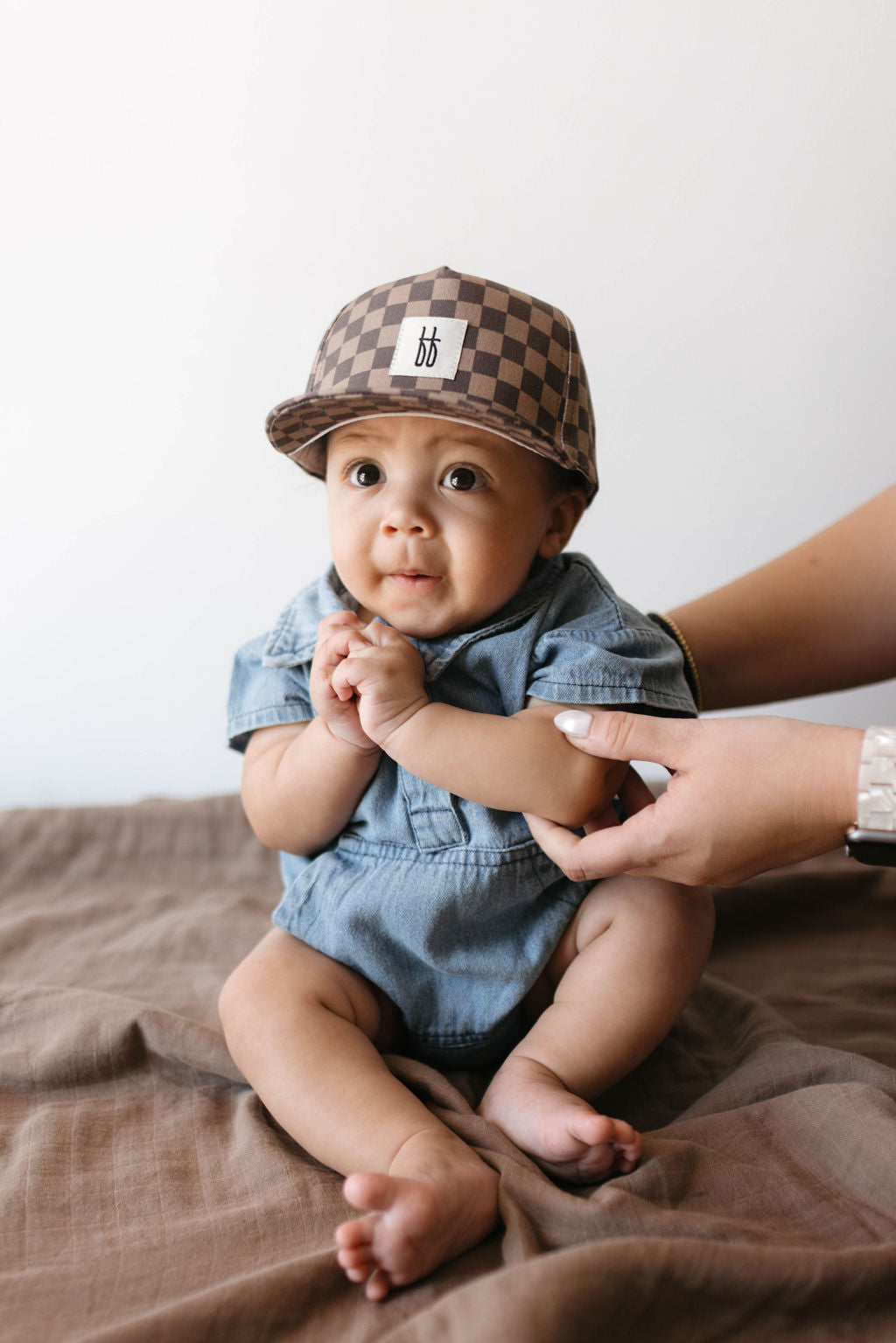 A baby, dressed in a chic Denim Romper from Forever French Baby and wearing a checkered brown cap, sits on a brown blanket. The baby's hands are clasped together, supported by an adult's hands from behind. The background is plain white.