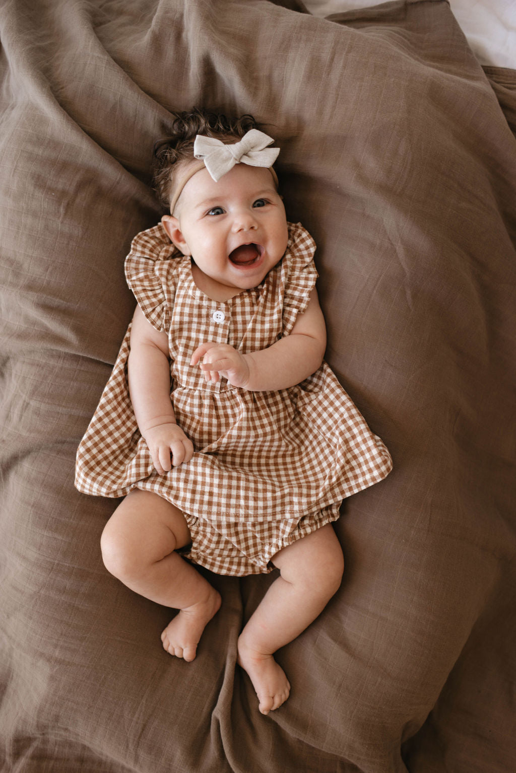 A smiling baby wearing the Honey Gingham Bloomer Set and a white bow headband lies on brown bedding. The baby, part of the Forever French Baby collection, is positioned in the center of the image with arms and legs slightly raised.