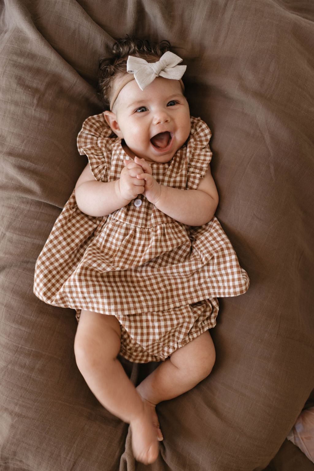 A joyful baby wearing the Gingham Bloomer Set in Honey from Forever French Baby and a cream-colored bow headband is lying on brown bedding. The baby has a big smile, hands clasped together, and appears to be laughing or giggling.