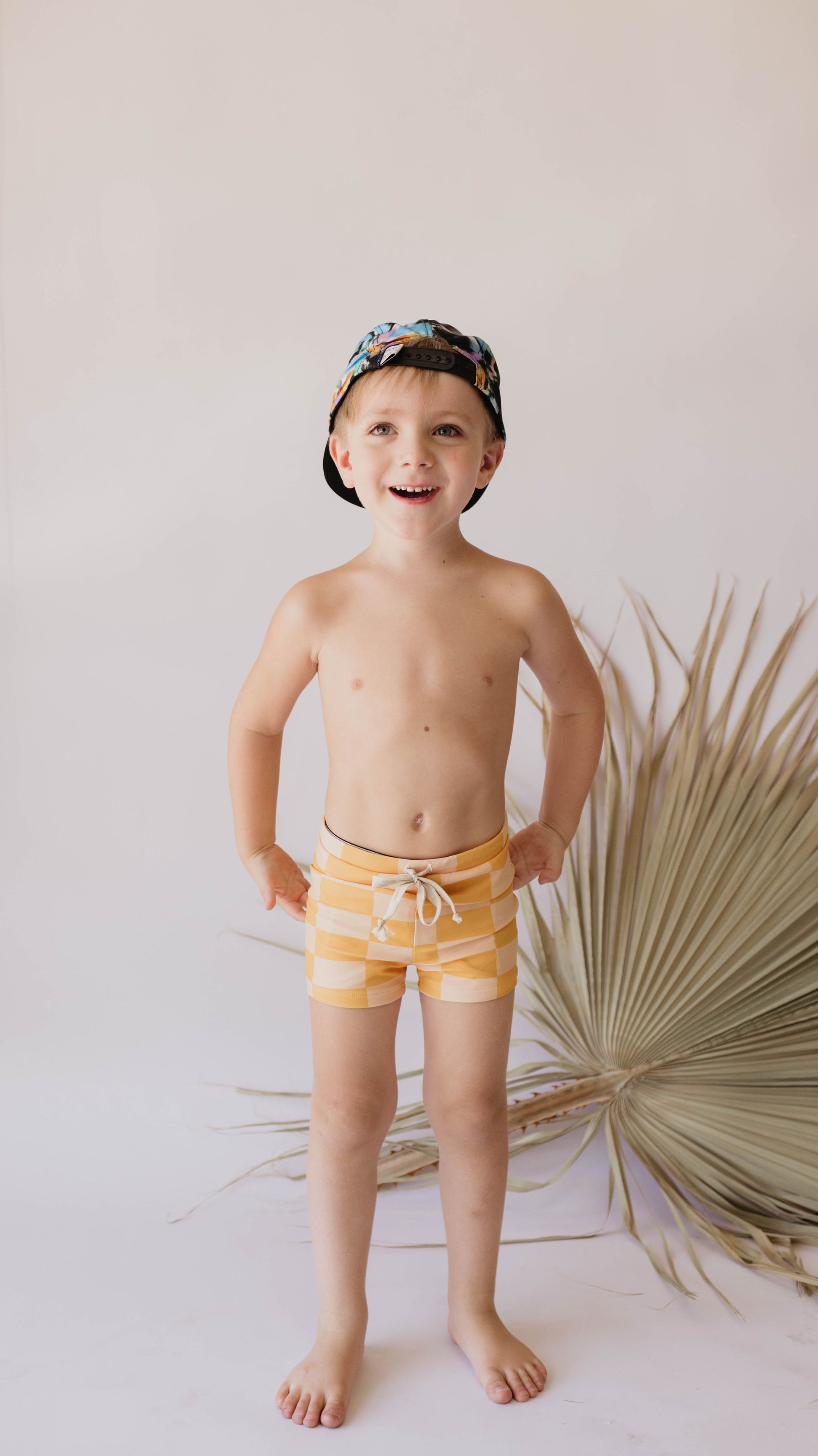 A young child stands confidently in front of a neutral background adorned with a dried palm leaf decoration. Sporting the vibrant Euro Swim Short | Sunny Checker from forever french baby, the child is dressed in yellow and white striped shorts that offer UPF 50+ protection, and wears a colorful cap. With hands on hips, the child beams joyfully.