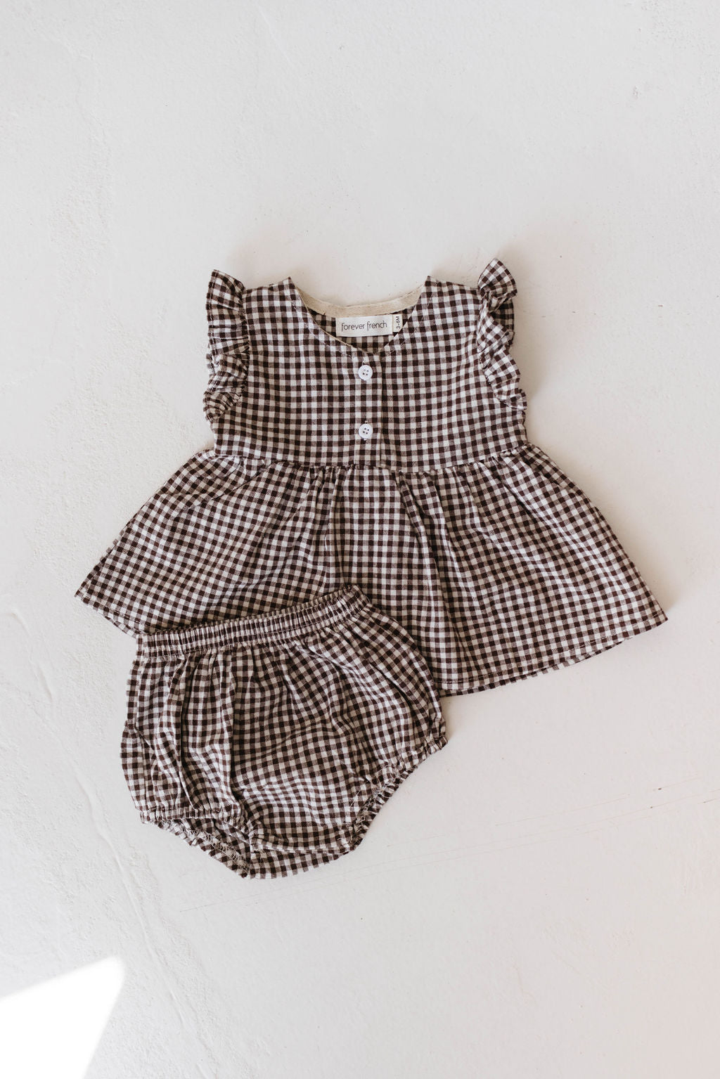 The Gingham Bloomer Set | Chocolate Chai by forever french baby includes a sleeveless brown and white gingham print dress with ruffled shoulders and two front buttons, paired with matching bloomers. The "Brown Tartan" fabric label is visible inside the 100% cotton dress. Both items are elegantly displayed on a plain surface.