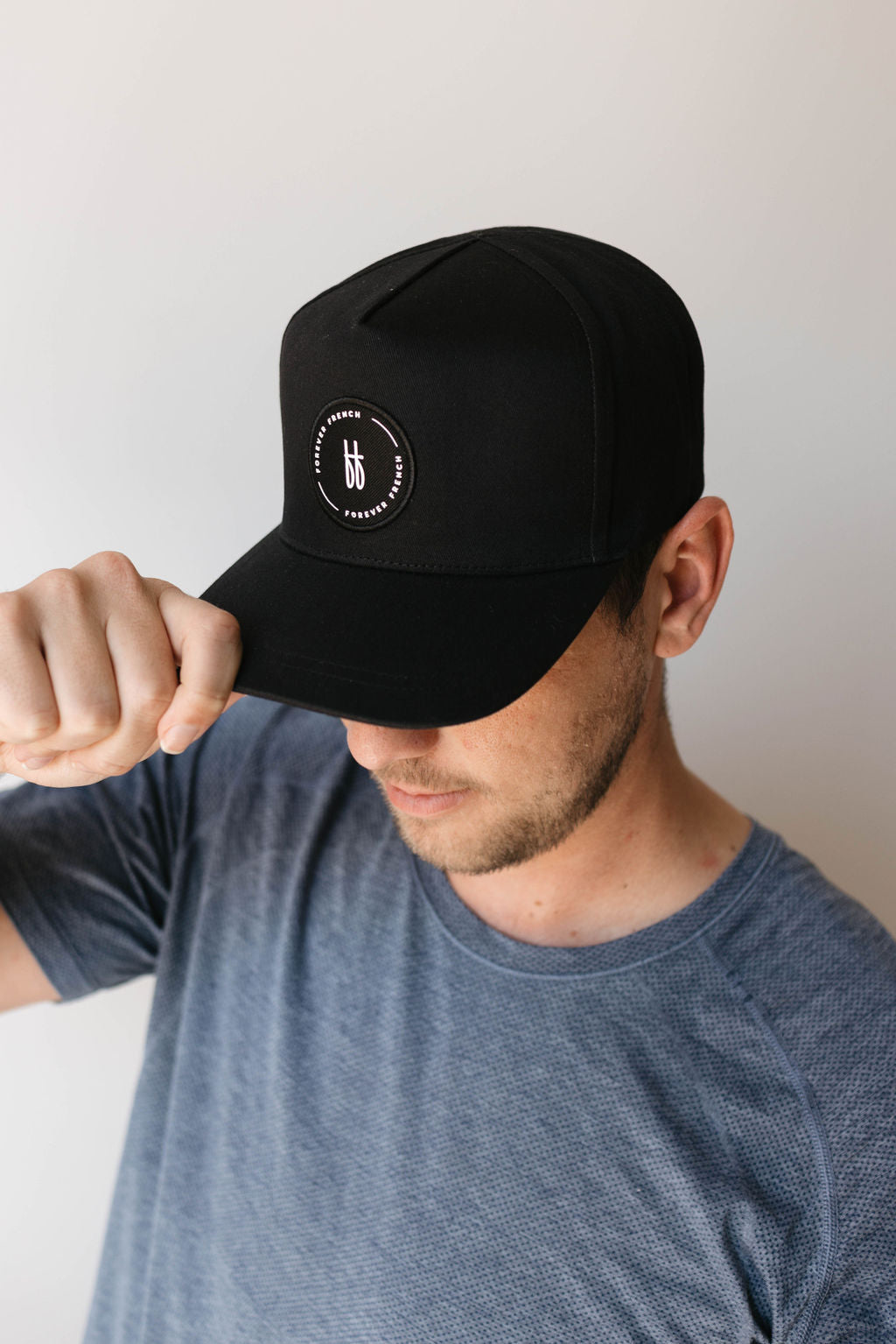 A person wearing a gray t-shirt is tilting the brim of their black Adult Trucker Hat by forever french baby, which features a logo with the letters "bf." The individual is looking down, and their face is partially obscured by the hat. The background is plain and light-colored—perfect for spot cleaning with mild detergent and warm water.