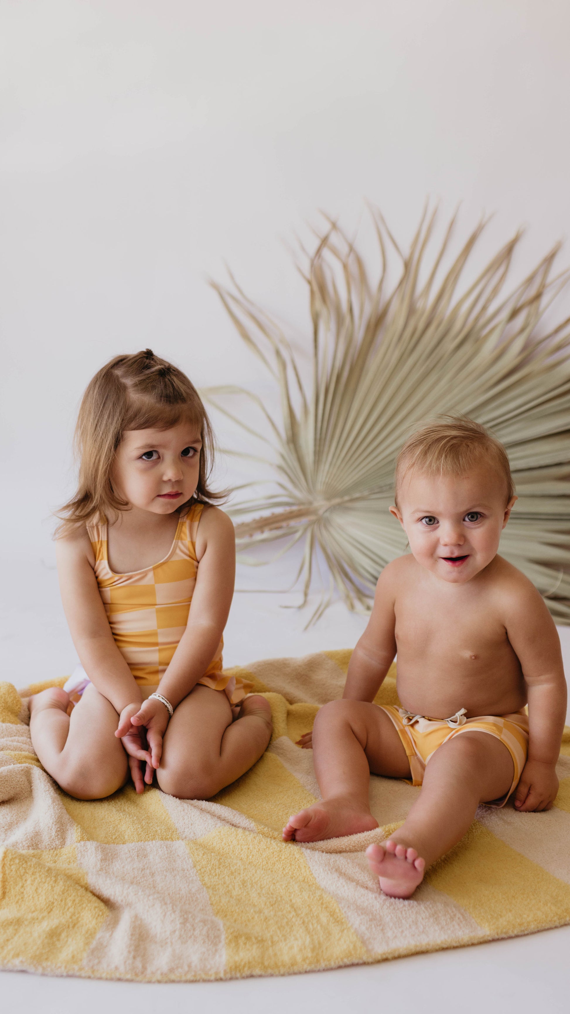 Two young children sit next to each other on a soft yellow blanket. The child on the left, in a forever french baby Girl Sleeveless Swimsuit | Sunny Checker, has shoulder-length brown hair. The child on the right, wearing yellow shorts, smiles widely. A large dried palm leaf is in the background.