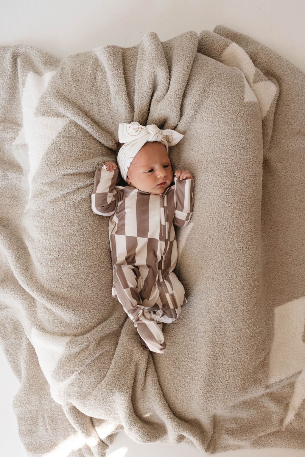 A newborn baby lies on a soft, beige blanket, wearing Forever French Baby's Bamboo Zip Pajamas in the Tile Stack design. The baby is also outfitted with matching mittens and a headband adorned with a bow. The plush blanket and the luxurious premium bamboo fabric create a cozy nest around the baby.