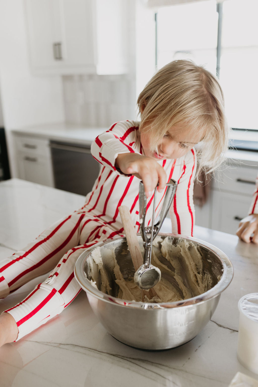 A young child wearing the Claus Bamboo Zip Pajamas by forever french baby sits on a kitchen counter, using an ice cream scoop to mix dough in a large metal bowl. Sunlight streams through a window, illuminating the modern kitchen setting and highlighting the comfort of their breathable attire.