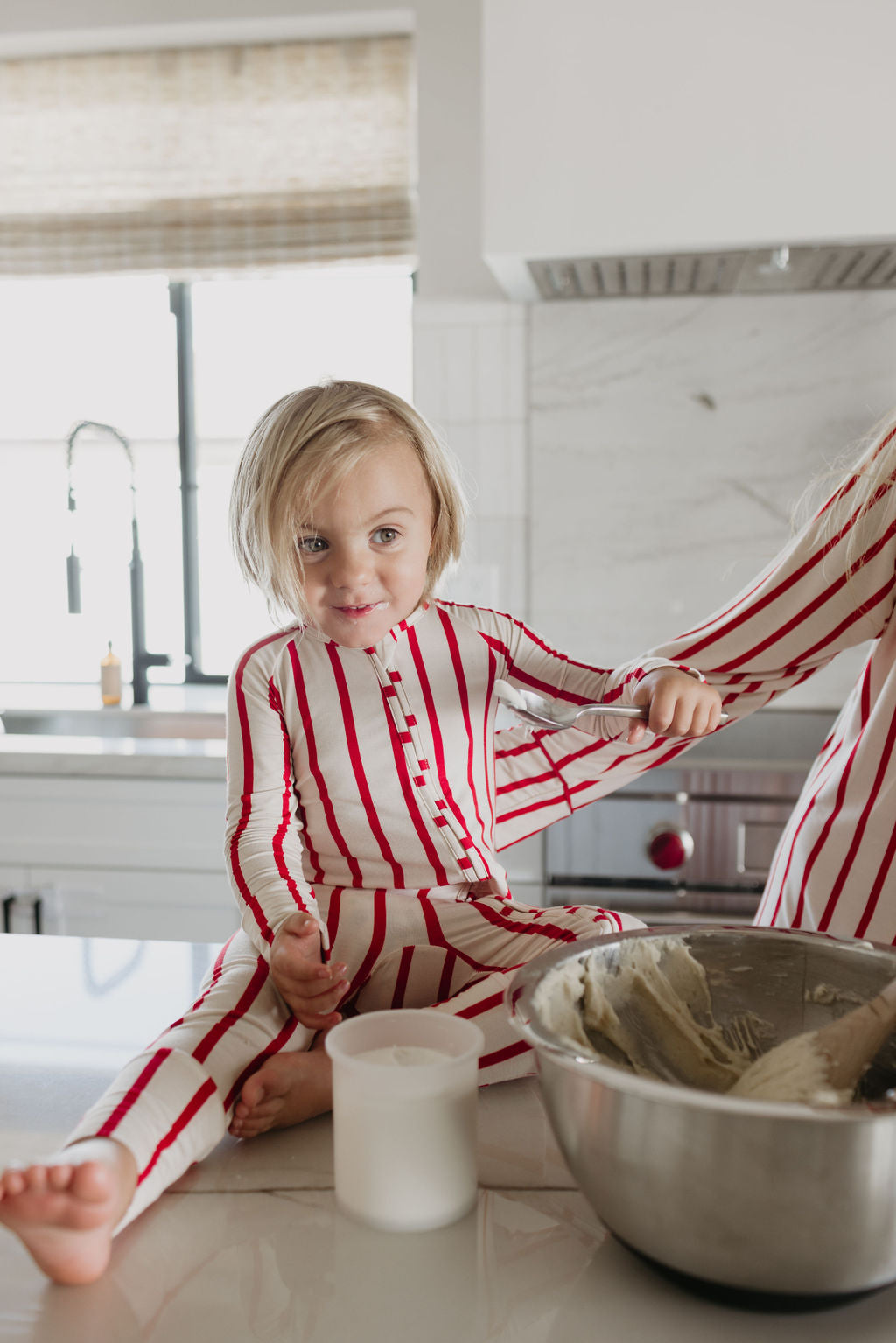 A child dressed in the Claus Bamboo Zip Pajamas by forever french baby sits on a kitchen counter beside a mixing bowl filled with dough. They are smiling and holding someone's hand, enjoying a cheerful moment in the bright kitchen.