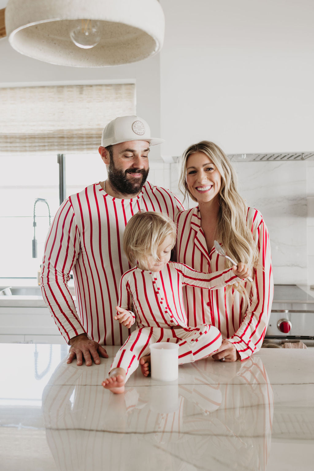 A family of three in matching red and white striped pajamas is gathered by a kitchen counter. The father sports a cap, the mother beams in her "Women's Bamboo Sleeping Dress | the Claus" by forever french baby, and their young child sits on the counter holding a mug. The kitchen is bright and modern.