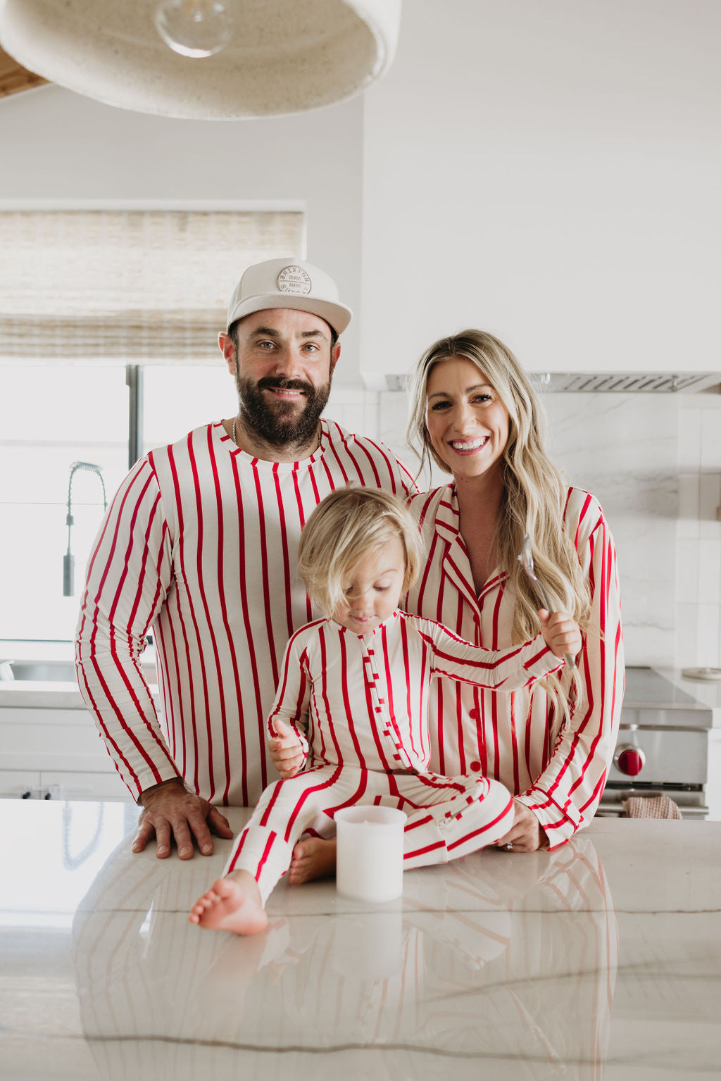 A smiling family stands in an eco-friendly kitchen, all wearing matching "the Claus" pajamas from forever french baby. The child sits on the counter, held by the woman dressed in a Women's Bamboo Sleeping Dress, while the man stands beside them. A hypo-allergenic candle is placed on the countertop in front of the child.