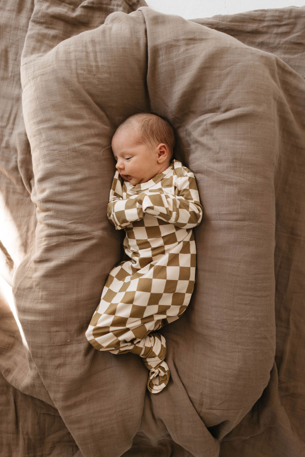 A newborn baby sleeps peacefully on a soft, beige blanket, dressed in the "Bamboo Knotted Gown | Olive Checkerboard" by forever french baby. With arms gently crossed and eyes closed, the serene setting highlights the coziness and comfort surrounding the infant.