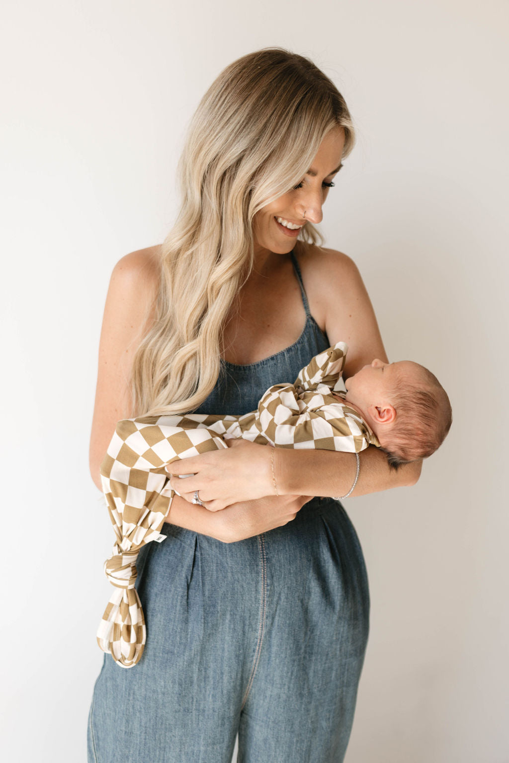 A woman with long blonde hair, wearing a sleeveless denim jumpsuit, is holding a newborn baby wrapped in the "Olive Checkerboard" Bamboo Knotted Gown by forever french baby. She is looking down and smiling at the baby. The background is plain and light. The baby's gown is part of their hypo-allergenic bamboo clothing line.