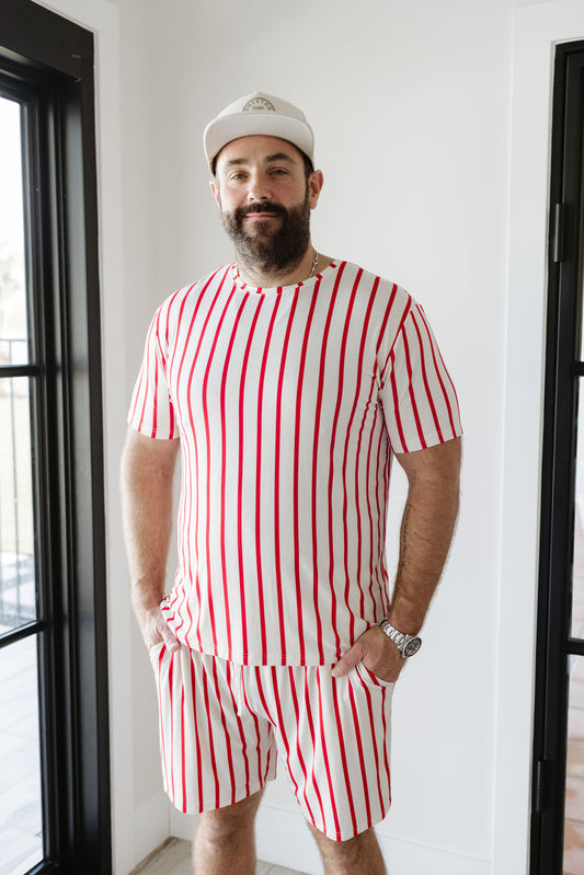 A man stands indoors wearing the "the Claus" short sleeve men's bamboo pajamas from Forever French Baby, featuring a matching red and white vertical striped shirt and shorts. He sports a light-colored cap and watch, with his hands in his pockets, all framed by black-framed windows in the background.