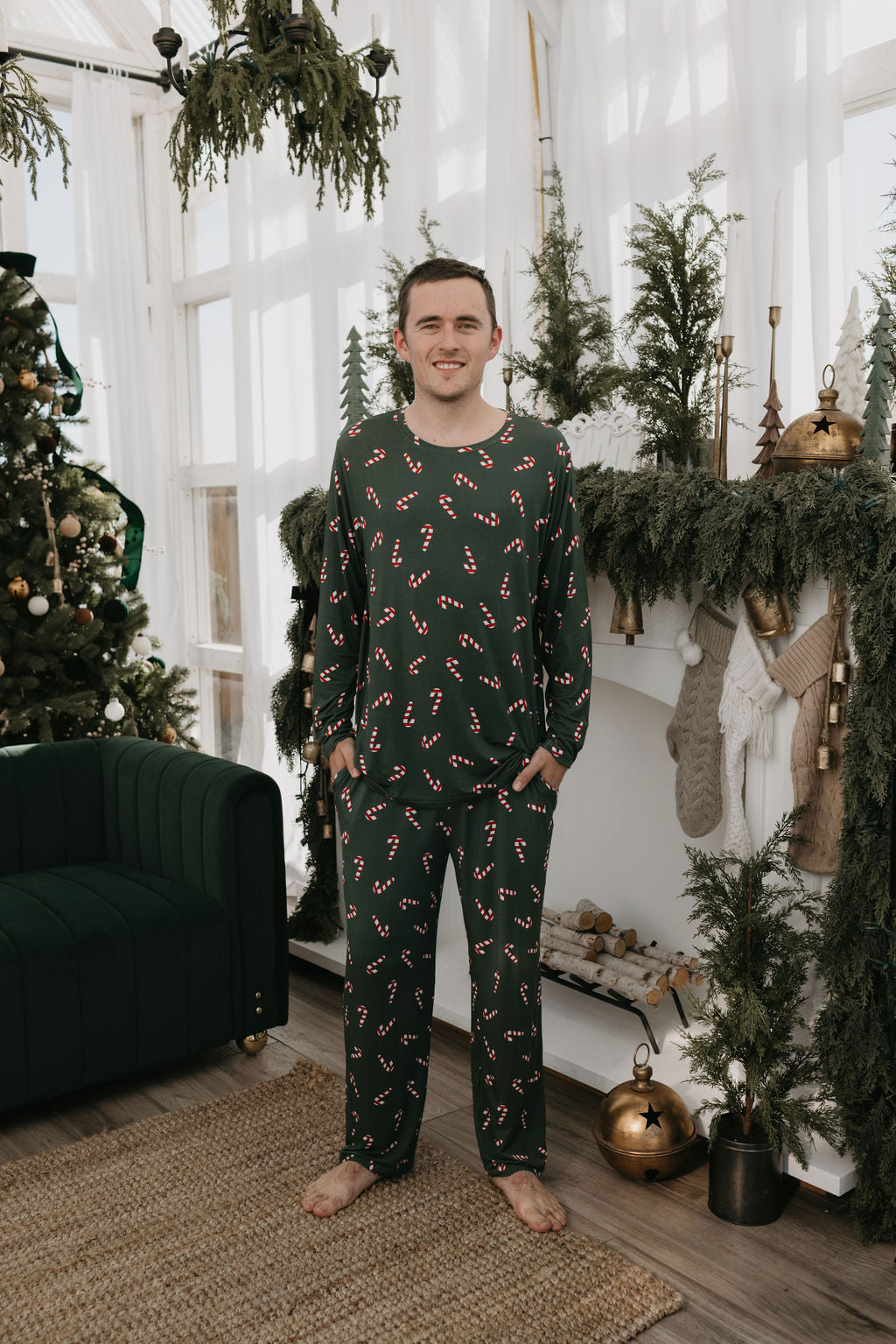 A person stands barefoot in a cozy living room, wearing the Men's Bamboo Pajamas from the Candy Cane Lane collection by forever french baby. The room is adorned with a Christmas tree, hanging stockings, and festive greenery, creating the perfect holiday backdrop.