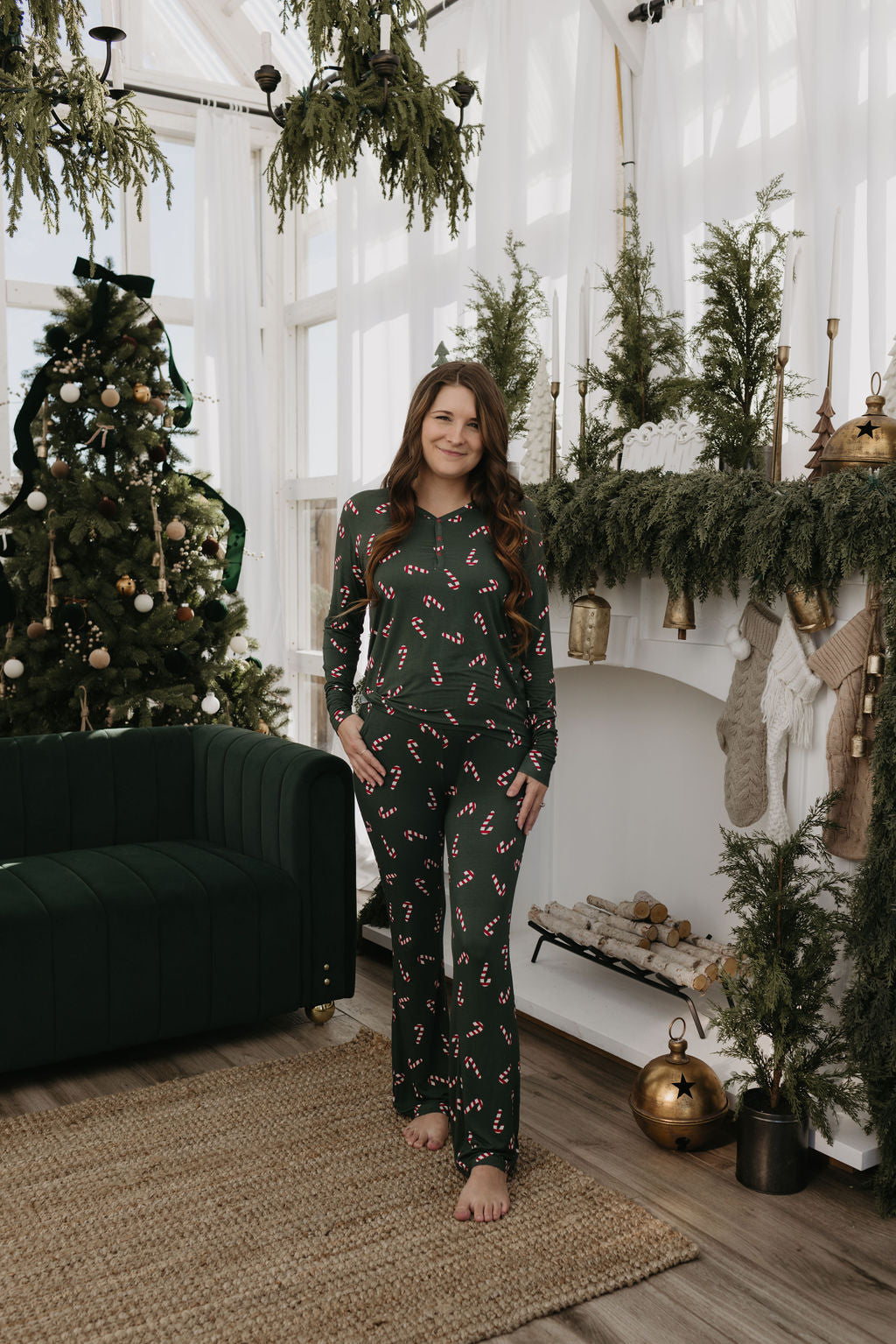 A woman dressed in Women's Bamboo Pajamas | Candy Cane Lane by forever french baby stands in a festive living room. The room showcases a decorated Christmas tree, a green couch, and a fireplace adorned with stockings and greenery. The cozy, holiday-themed atmosphere is ideal for breastfeeding moms seeking comfort.