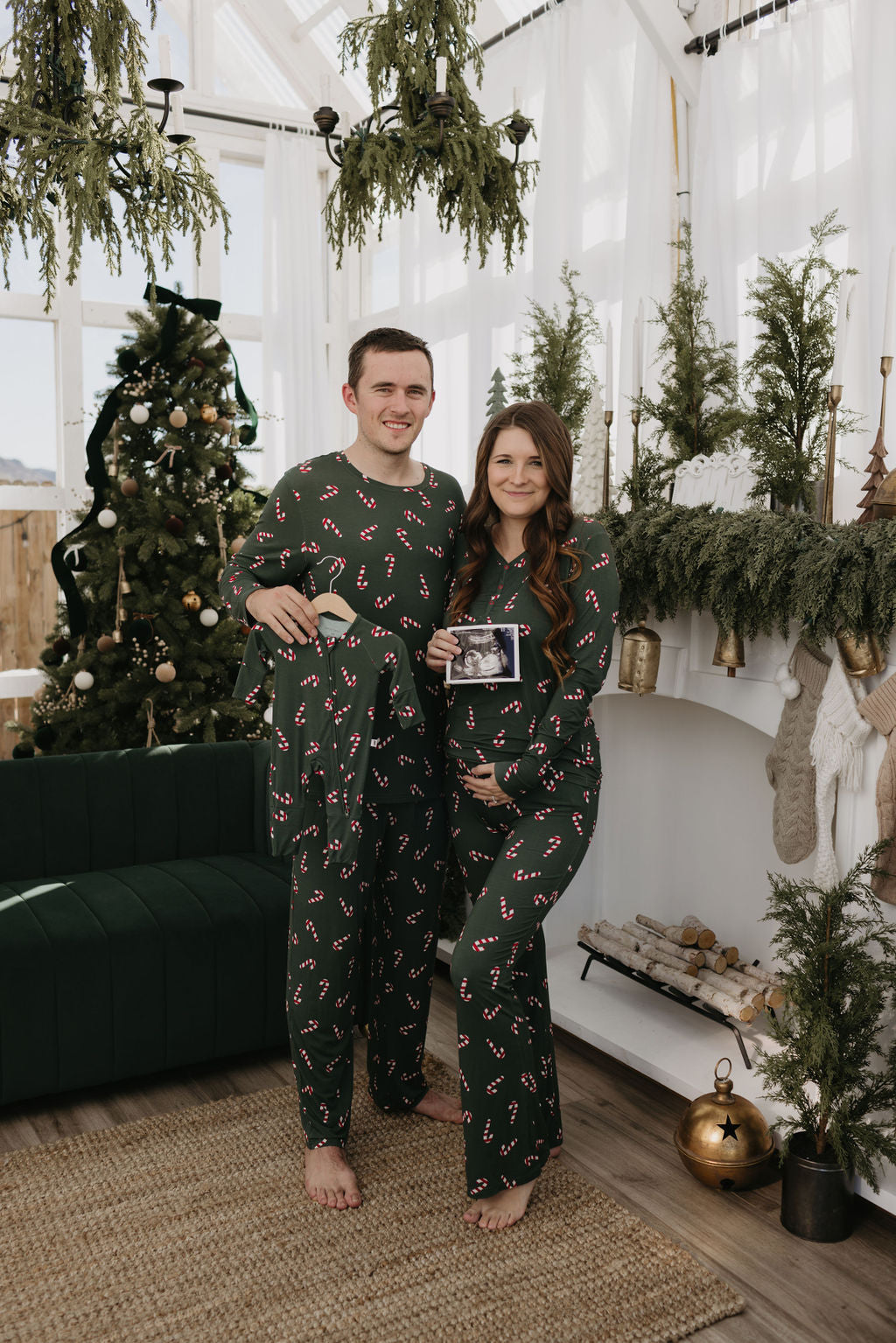 A couple dressed in matching pajamas from the forever french baby's Candy Cane Lane collection stands in a festive living room. The woman holds a sonogram, while the man showcases a tiny outfit. A decorated tree and greenery garlands enhance the holiday ambiance.