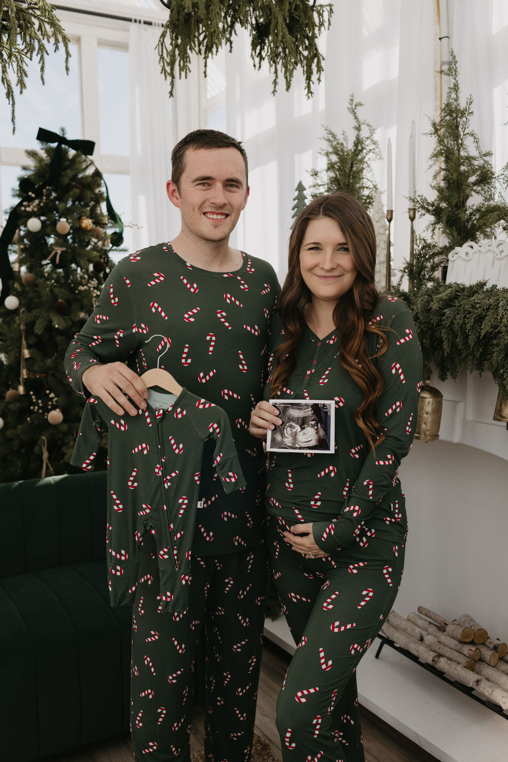 A couple wearing matching forever french baby Women's Bamboo Pajamas in the Candy Cane Lane design stands in a festive room adorned with a decorated Christmas tree. The woman, smiling, holds an ultrasound photo and cradles her belly, while the man lifts a tiny matching pajama set designed for their soon-to-arrive little one to enjoy cozy family moments.