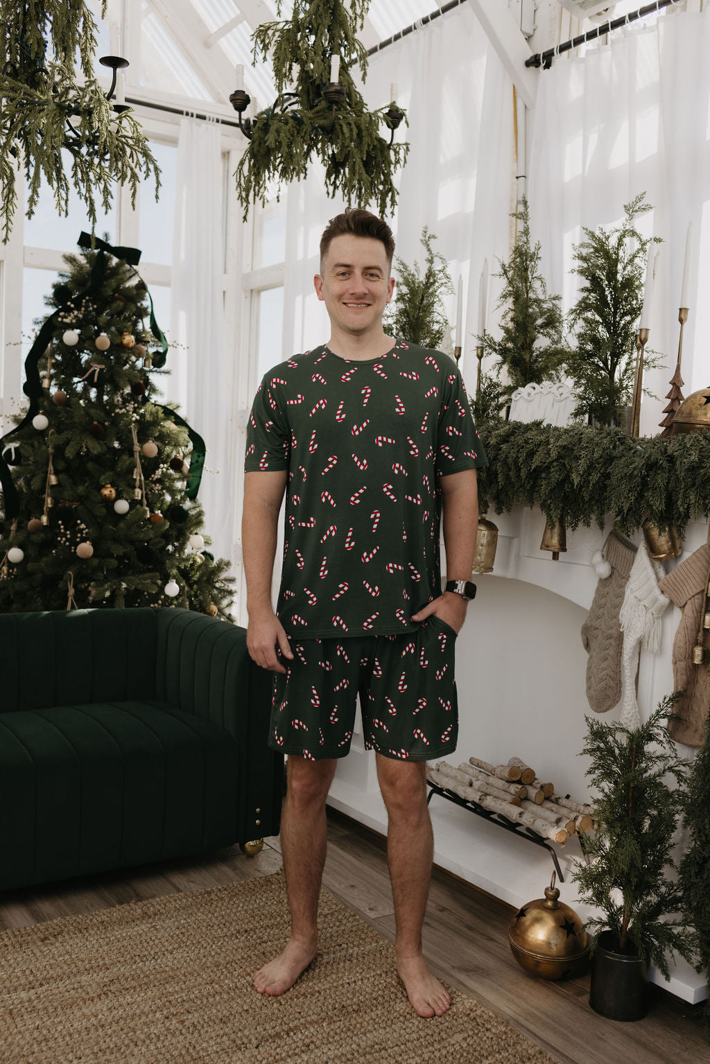 In a living room festively adorned with a Christmas tree, garlands, and a fireplace accentuated by greenery and ornaments, someone is wearing the comfortable and breathable hypo-allergenic Short Sleeve Men's Bamboo Pajamas in the Candy Cane Lane pattern by forever french baby.