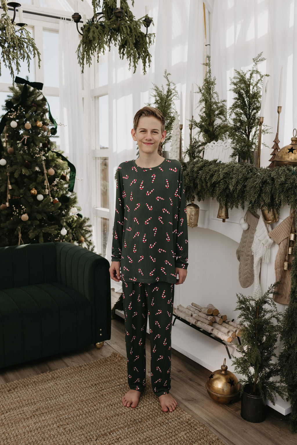 A joyful child wearing dark green pajamas from forever french baby's Candy Cane Lane collection stands in a festive living room adorned for Christmas, surrounded by a tree, garlands, and stockings by the fireplace.