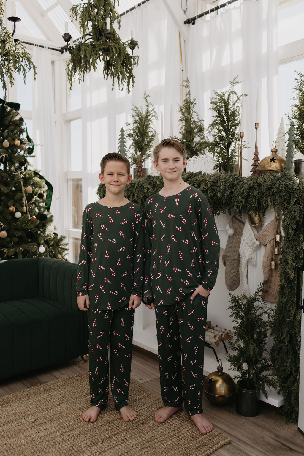 Two children wearing matching Pre-Teen Straight Leg Bamboo Pajamas from the Candy Cane Lane collection by forever french baby stand in a festive living room. A Christmas tree and a mantelpiece decorated with stockings and greenery can be seen in the background, as natural light filters through large windows.