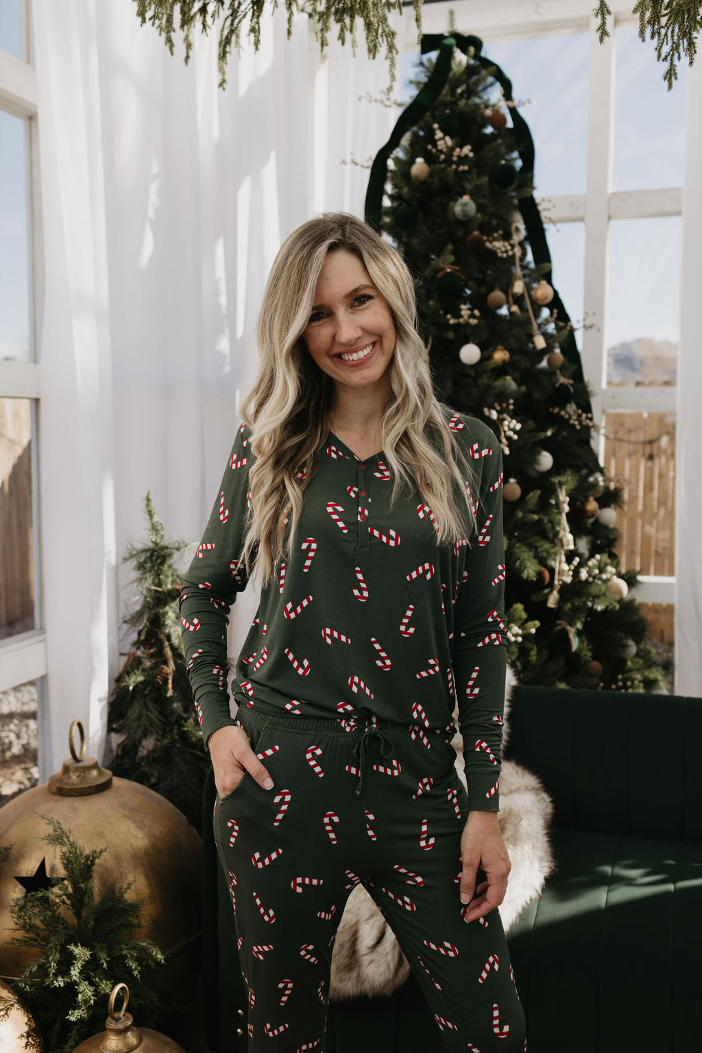 A woman in the Women’s Bamboo Pajamas | Candy Cane Lane by forever french baby stands smiling in front of a decorated Christmas tree. The room features festive decorations, including large gold ornaments and greenery, creating a cozy atmosphere ideal for breastfeeding moms seeking comfort and style.