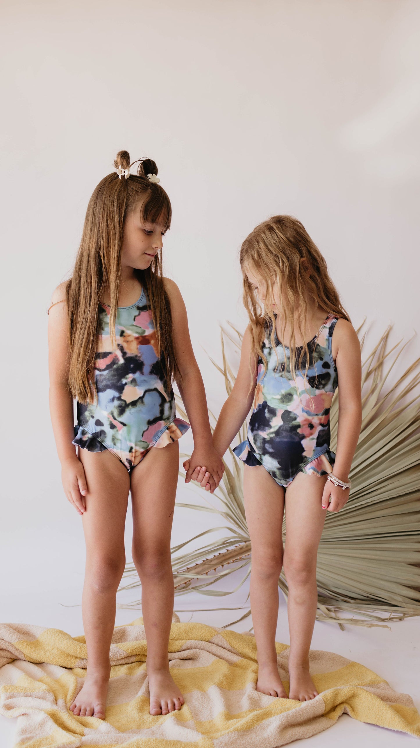 Two young children with long hair, wearing matching colorful Girl One Piece Swimsuits | Charli from forever french baby, hold hands while standing on a towel. They are in front of a dried palm leaf backdrop and both look downwards, sharing a moment that subtly promotes Autism awareness.