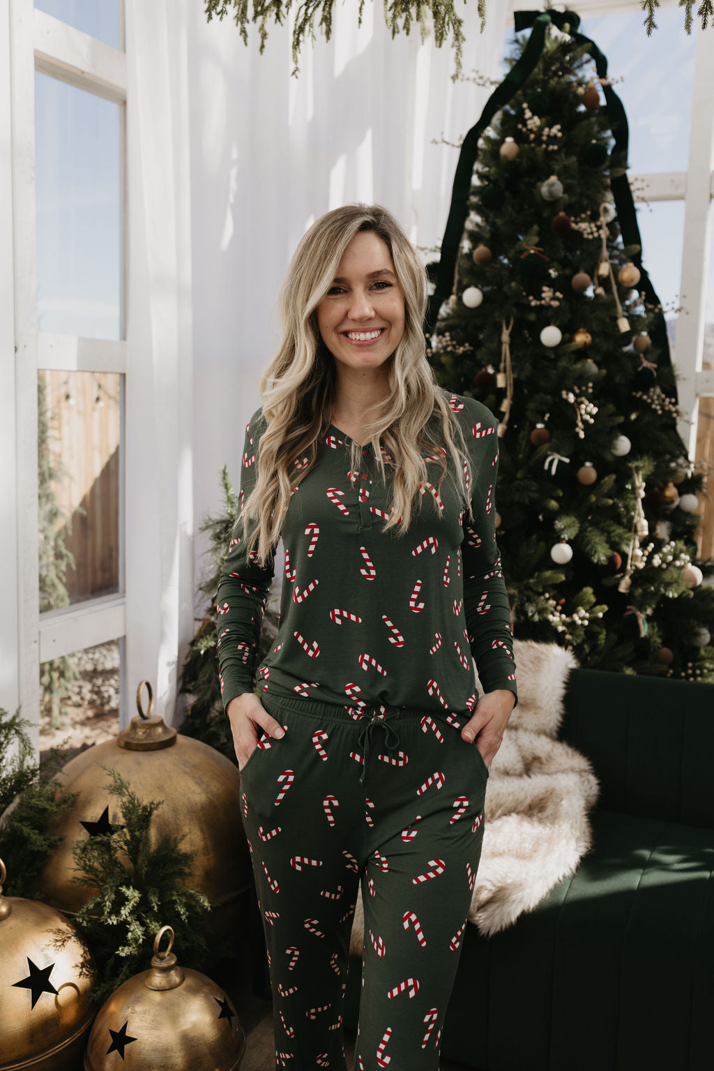 A woman wearing Women's Bamboo Pajamas from the Candy Cane Lane collection by forever french baby stands in front of a decorated Christmas tree and large gold ornaments. She smiles with her hands in her pockets, enjoying the festive, warmly lit room—offering perfect comfort for breastfeeding moms.