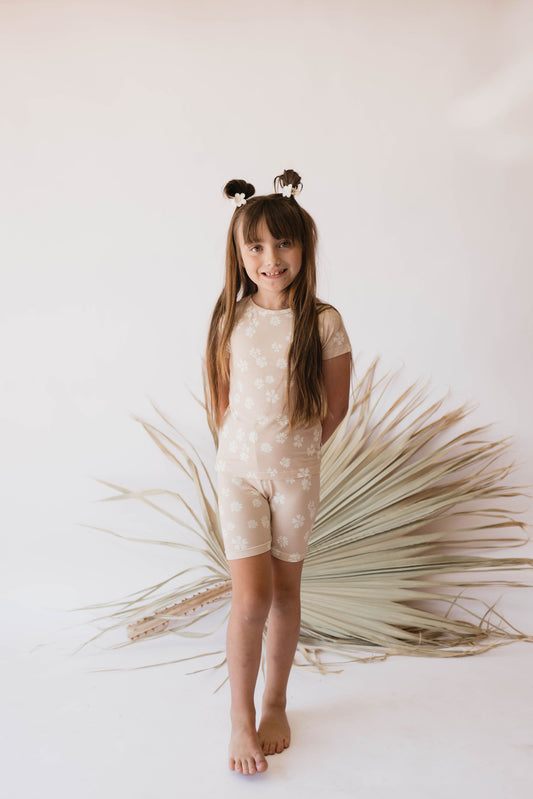 A young girl with long hair stands barefoot in front of a white backdrop adorned with dried foliage. She is wearing the hypo-allergenic "Lazy Daisy | Bamboo Short Set" by forever french baby, featuring a beige outfit with a floral pattern and has her hands behind her back. Her two small hair buns on top of her head are accessorized with flower-like decorations.