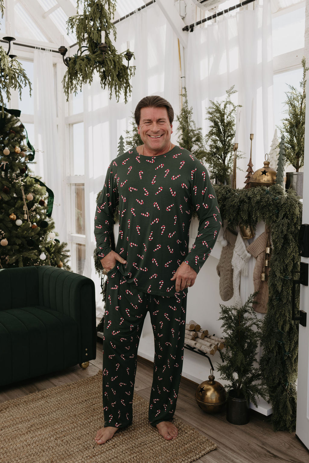 In a cozy room adorned with holiday decorations, a man is dressed in the Men's Bamboo Pajamas from the Candy Cane Lane collection by forever french baby. These hypo-allergenic pajamas, featuring green fabric decorated with red and white candy canes, enhance the charm of the scene. The festive atmosphere is completed by a Christmas tree, stockings, and greenery throughout the room.