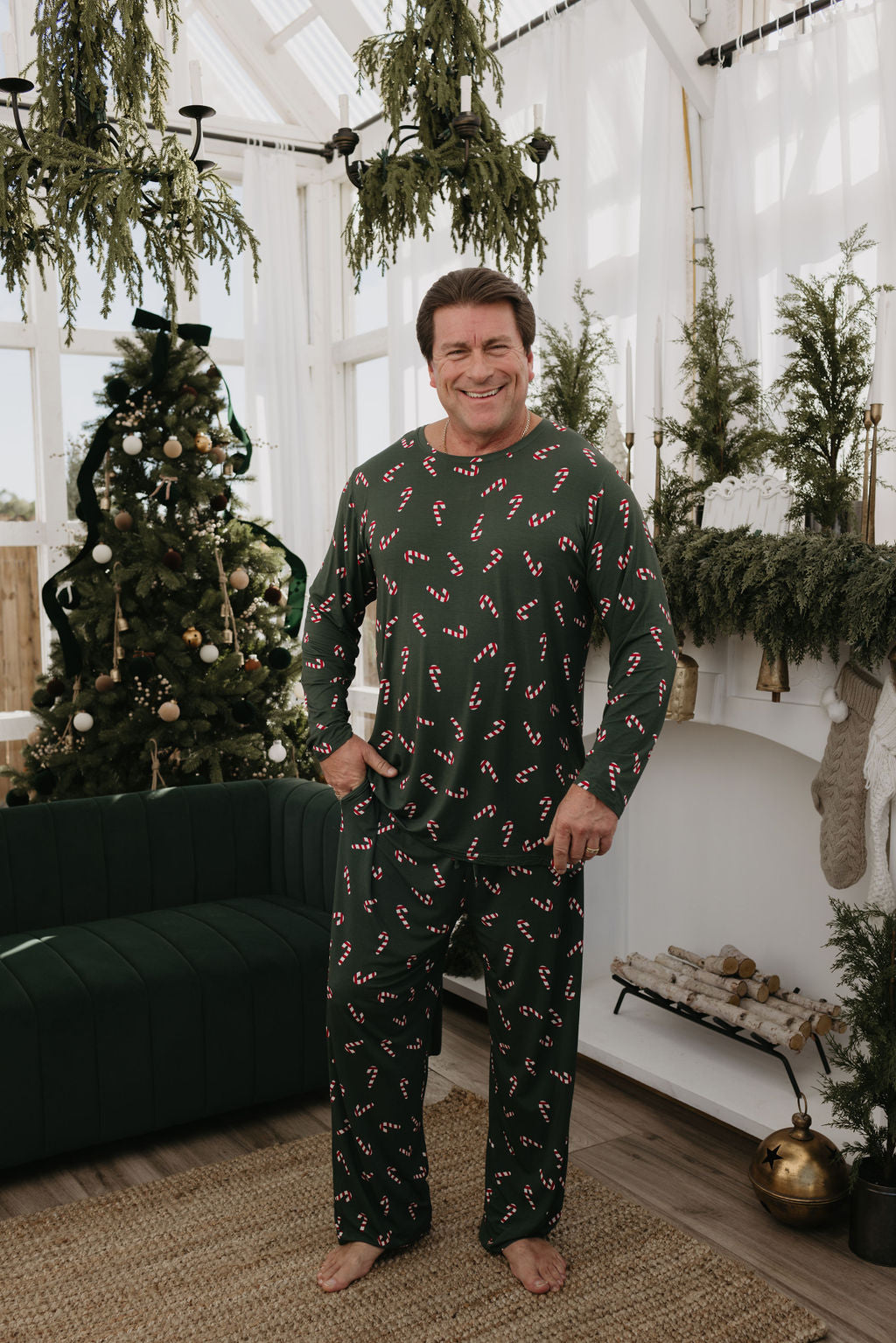 A man wearing Men's Bamboo Pajamas from the Candy Cane Lane collection by forever french baby stands in a festive room with a decorated Christmas tree and greenery on the fireplace. He is barefoot, smiling, and the room radiates a cozy holiday atmosphere.