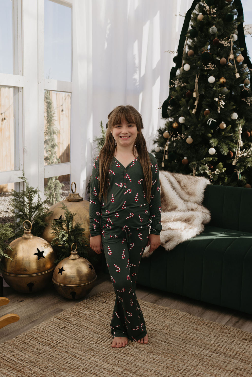 A young girl with long brown hair stands barefoot on a woven rug, wearing the Pre-Teen Flare Bamboo Pajamas from forever french baby in the Candy Cane Lane design. She smiles in a festive room highlighted by a decorated Christmas tree and large gold bells.