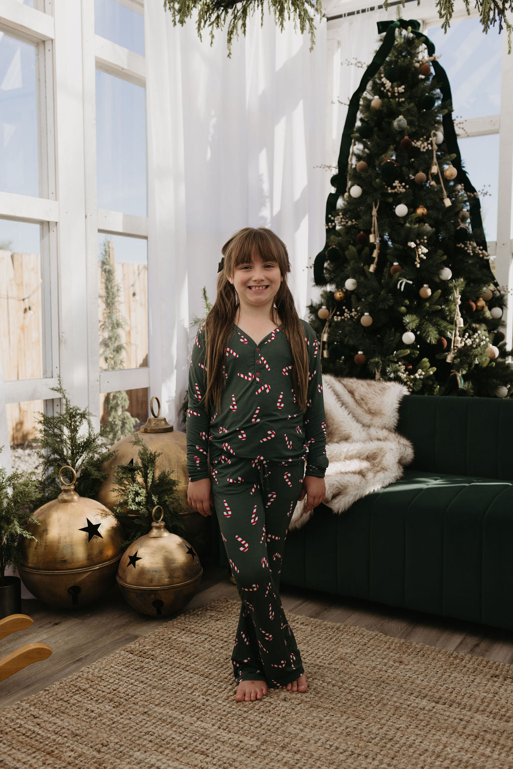 A young girl beams in front of a Christmas tree, dressed in forever french baby's Pre-Teen Flare Bamboo Pajamas from the Candy Cane Lane collection. The room radiates festive cheer with large gold ornaments and greenery. She stands barefoot on a woven rug, embodying the essence of holiday comfort and joy.