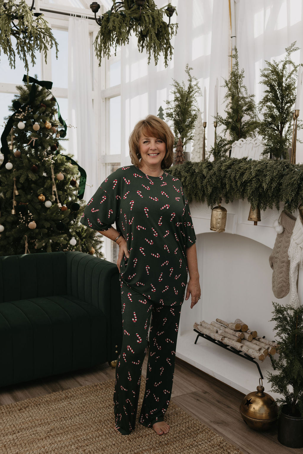 Wearing the Short Sleeve Women's Bamboo Pajamas from forever french baby's Candy Cane Lane collection, a woman stands in a cozy, festively decorated living room. The bamboo pajamas offer both comfort and style, perfectly complementing the Christmas tree, garlands, and stocking-adorned fireplace as natural light streams through large windows.