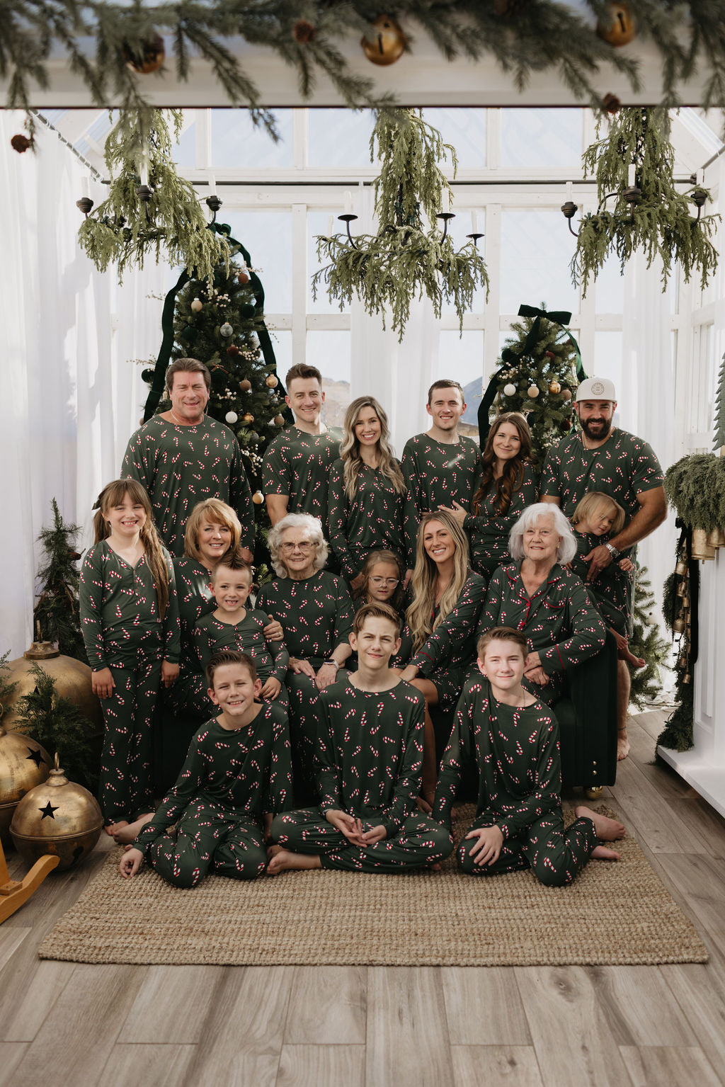 A large family dressed in matching pajamas from the Candy Cane Lane collection by forever french baby poses for a holiday photo. Gathered indoors amidst decorated Christmas trees, with greenery and ornaments framing the scene, a dog sits contentedly between two family members in the back row.