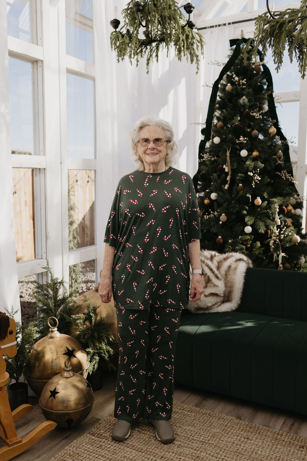 An elderly woman stands in front of a Christmas tree, wearing the Short Sleeve Women's Bamboo Pajamas from forever french baby, featuring the Candy Cane Lane pattern. The room is decorated for the holidays with festive ornaments and greenery.