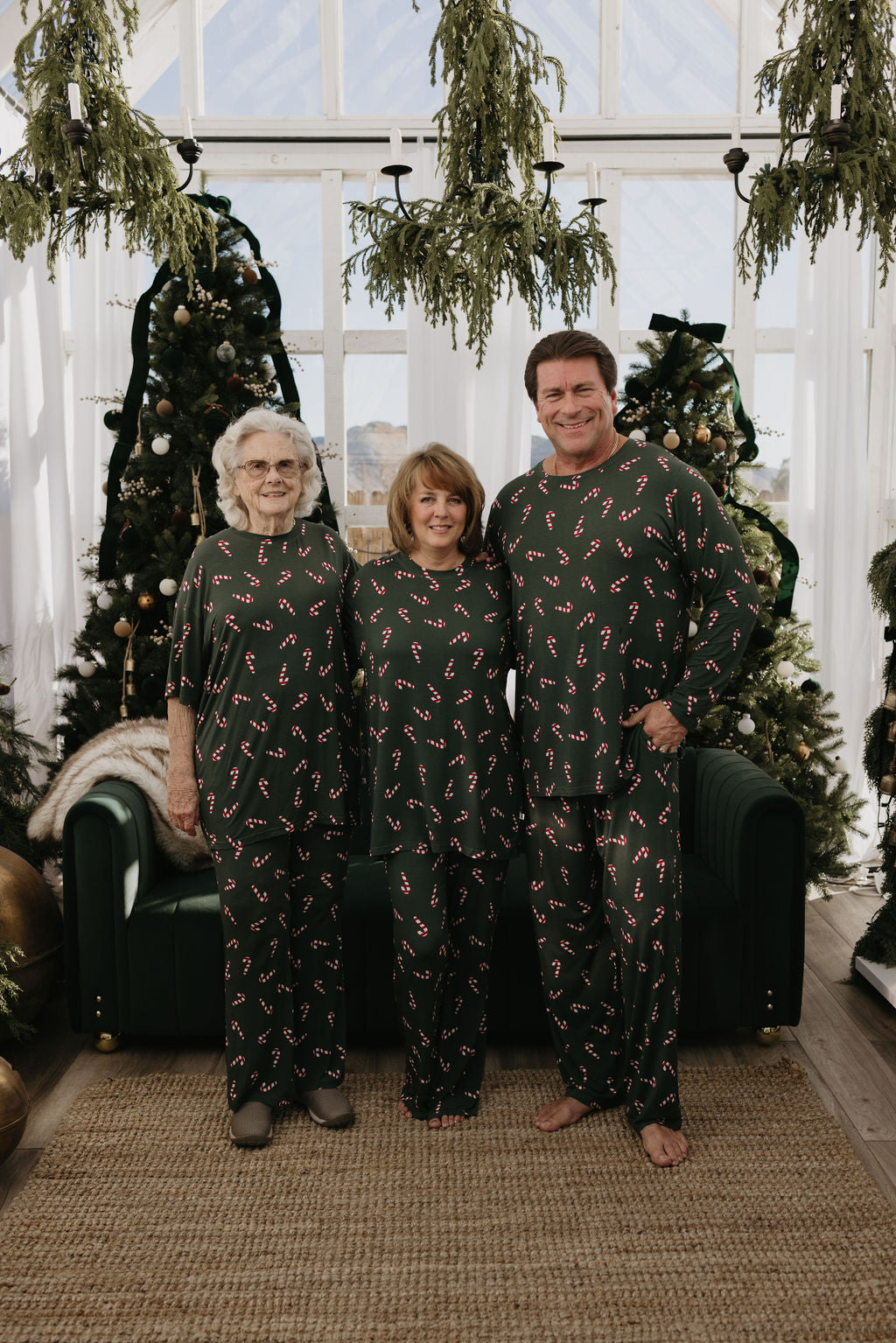 Three people don matching Men's Bamboo Pajamas from the Candy Cane Lane collection by forever french baby, featuring festive patterns, in a room beautifully decorated with Christmas trees and greenery. They are smiling and clearly enjoying the holiday magic.