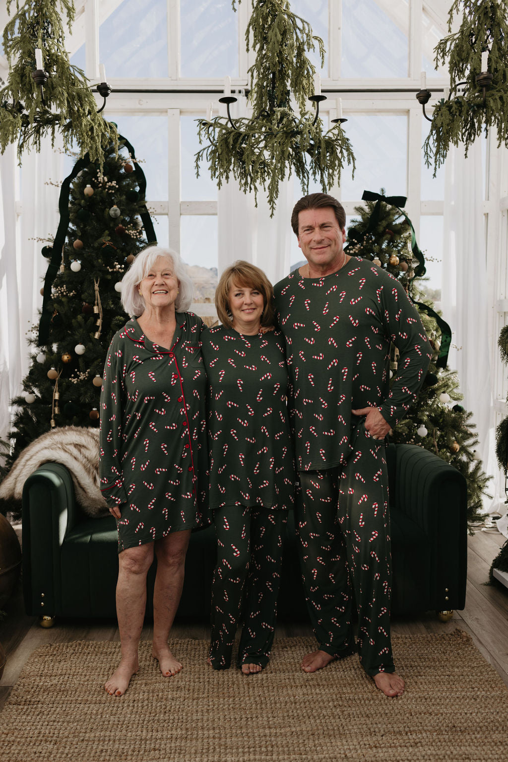 Three people wearing matching green Men's Bamboo Pajamas from the forever french baby Candy Cane Lane collection, adorned with red candy cane patterns, stand together smiling and barefoot by the Christmas tree. The indoor setting features large windows and decorative greenery, capturing the holiday spirit.