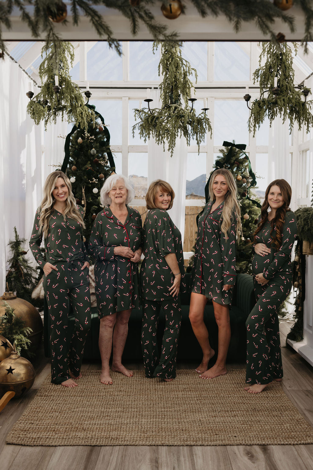 Five women in matching oversized "Short Sleeve Women's Bamboo Pajamas" from the Candy Cane Lane collection by forever french baby stand indoors. They smile and pose in front of decorated Christmas trees, surrounded by hanging greenery and ornaments. Natural light filters through a glass ceiling, illuminating their joyful gathering.