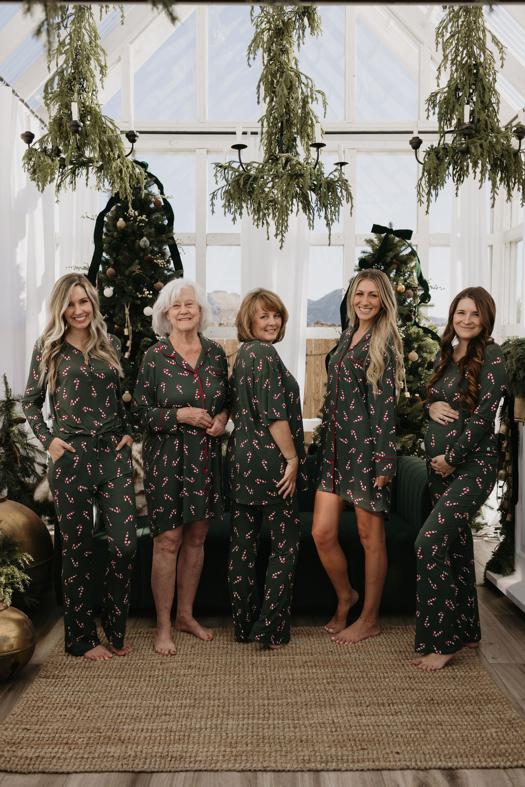 Five women of varying ages stand barefoot in matching "Women's Bamboo Sleeping Dresses | Candy Cane Lane" by forever french baby. They are smiling and posing in a festive room adorned with green garlands and a Christmas tree as the backdrop.