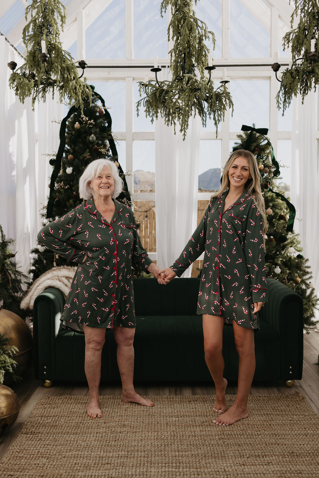 Two women wearing coordinated green, eco-friendly bamboo sleeping dresses from the "Candy Cane Lane" collection by forever french baby stand hand in hand, smiling in a festive indoor setting. They are surrounded by decorated Christmas trees and bathed in natural light streaming through tall windows.