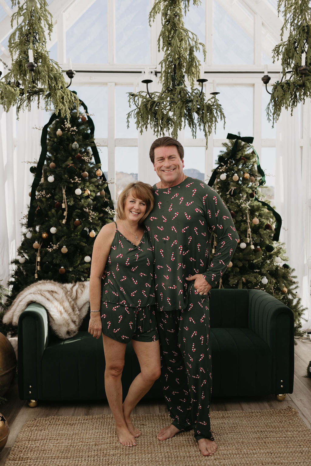 A smiling couple stands barefoot in front of a green couch, showcasing forever french baby's Candy Cane Lane collection with matching men's bamboo pajamas. Behind them are two decorated Christmas trees and hanging greenery, creating a bright, airy feel in the room with large windows.