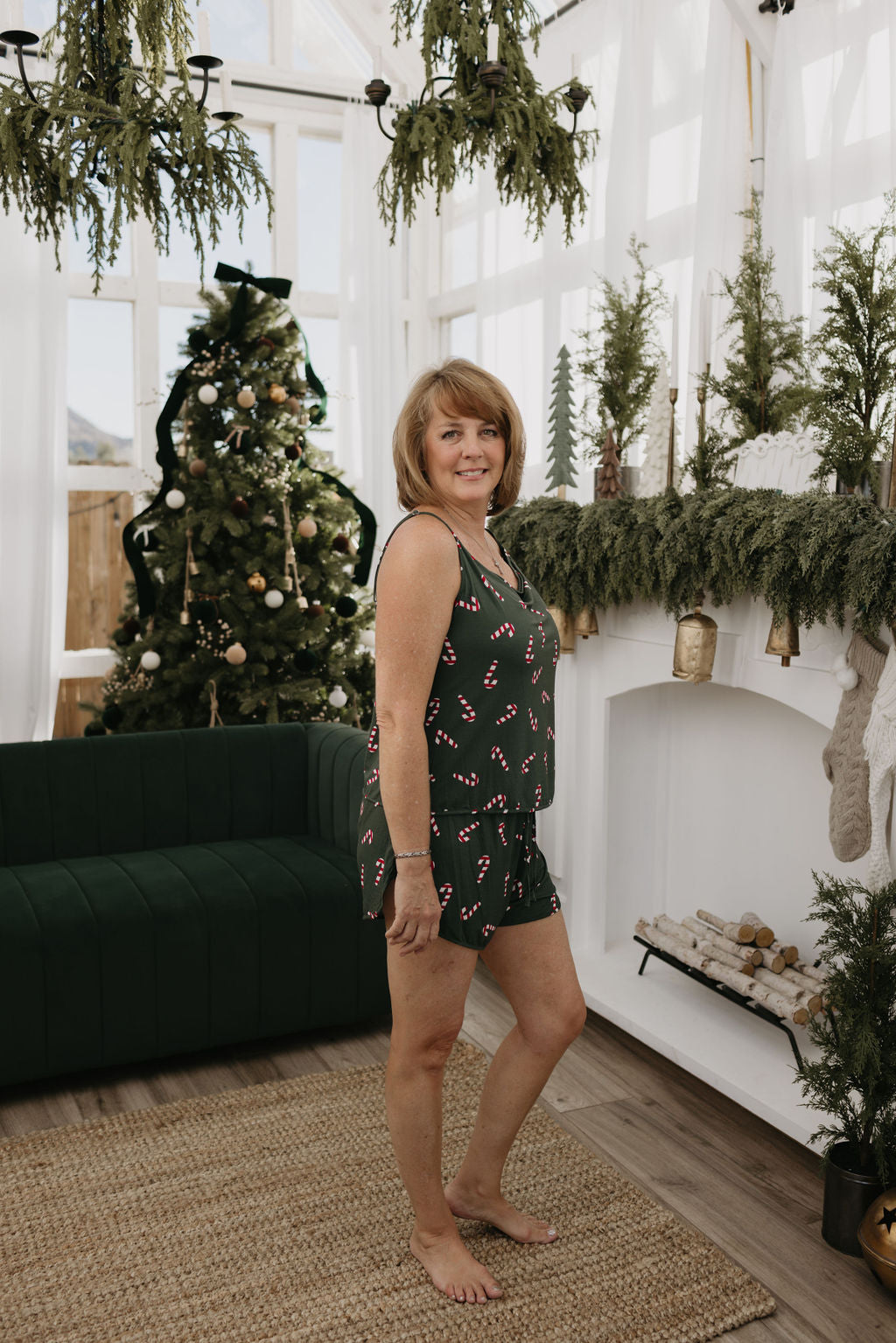 A woman wearing the forever french baby Cami Women's Bamboo Set in the Candy Cane Lane pattern, made from hypo-allergenic bamboo fabric, stands in a holiday-decorated living room with a green sofa, a Christmas tree, and a mantel adorned with greenery and stockings.