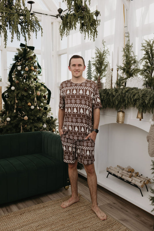 Wearing the "Short Sleeve Men's Bamboo Pajamas" in the Forever Fair Isle pattern by forever french baby, a person stands barefoot in a decorated room featuring a Christmas tree, garlands, and a white mantel with logs. The breathable fabric ensemble highlights their warm smile as they tuck their hands into pockets, fully embracing the cozy holiday spirit.