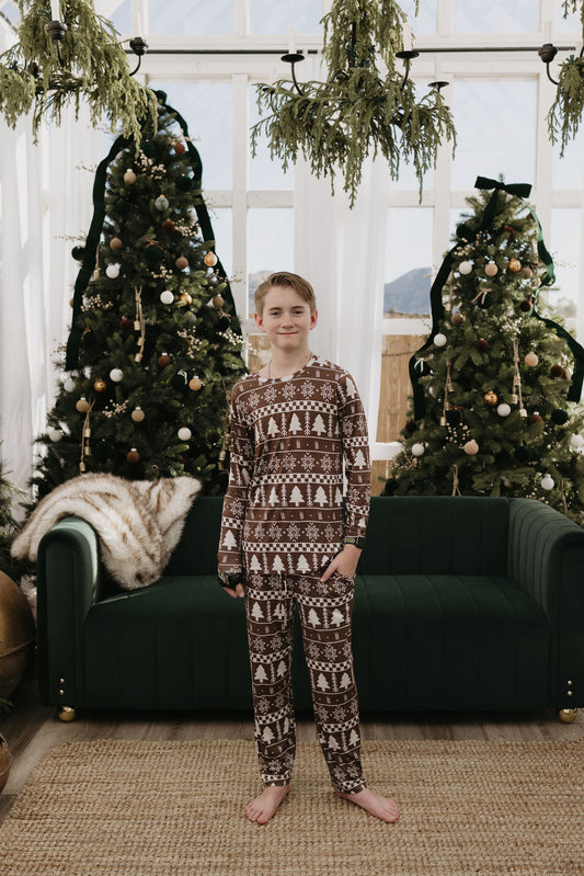 A young individual stands barefoot in front of a green sofa, dressed in festive Pre-Teen Straight Leg Bamboo Pajamas with a Fair Isle pattern from forever french baby. Behind them are Christmas trees adorned with lights and ornaments, situated in a bright and airy room bathed in natural light.