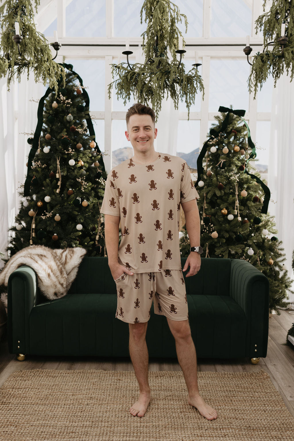 A person dressed in forever french baby's Men's Bamboo Short Pajamas in a gingerbread print stands barefoot on a rug before a dark green couch. Behind them, decorated Christmas trees and hanging greenery add to the festive atmosphere, while the breathable fabric guarantees comfort during celebrations.