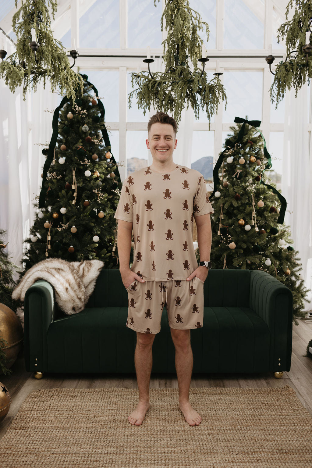 A person stands smiling in front of two festively adorned Christmas trees, wearing the Men's Bamboo Short Pajamas in a gingerbread pattern by forever french baby. The pajamas are crafted from breathable bamboo fabric. In the cozy room with large windows, a dark green couch and cheerful decorations complete the joyful atmosphere.